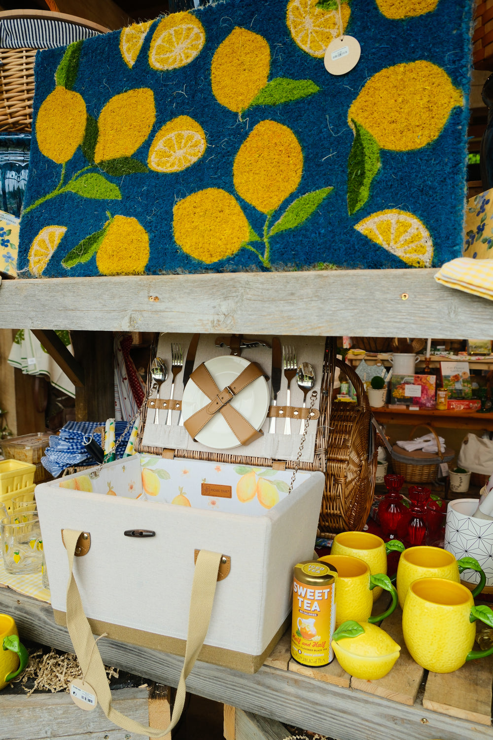 A rustic shop display featuring a blue doormat with a yellow lemon pattern, a white picnic basket with a wooden lid, a white cooler bag, and several yellow ceramic mugs on wooden shelves.